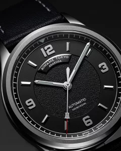 Close-up of black textured dial on an automatic wristwatch, featuring applied Arabic numerals, brushed hands, and minute track