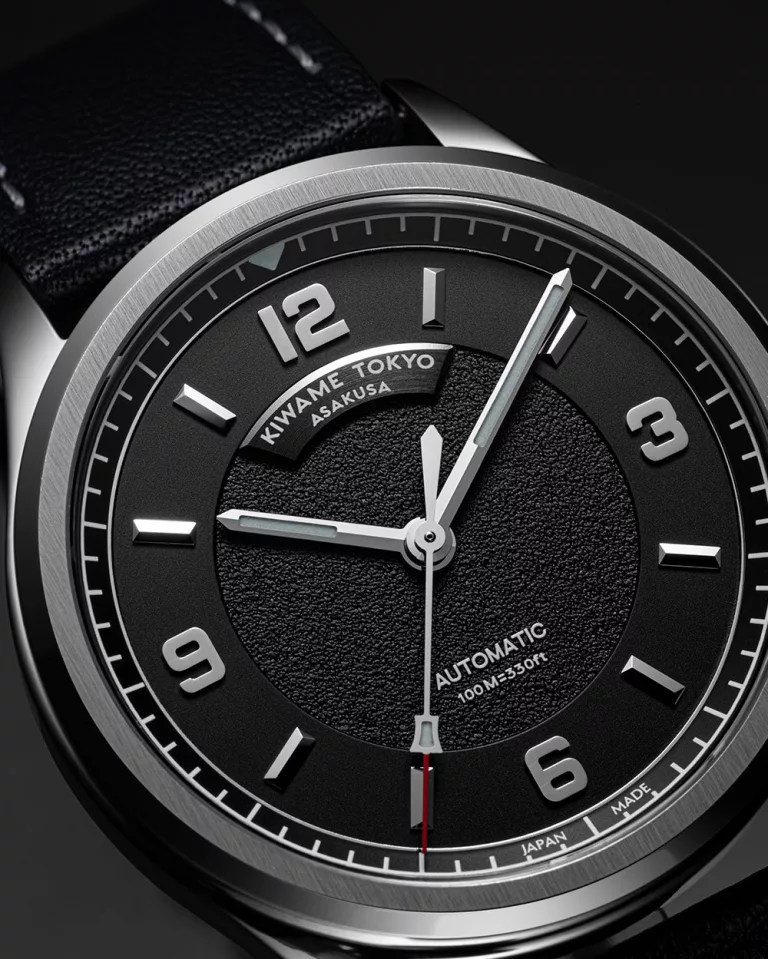 Close-up of black textured dial on an automatic wristwatch, featuring applied Arabic numerals, brushed hands, and minute track