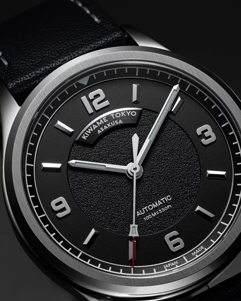 Close-up of black textured dial on an automatic wristwatch, featuring applied Arabic numerals, brushed hands, and minute track