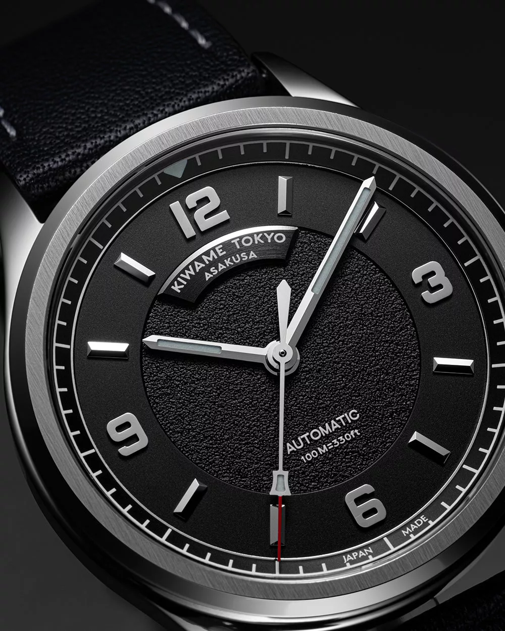 Close-up of black textured dial on an automatic wristwatch, featuring applied Arabic numerals, brushed hands, and minute track