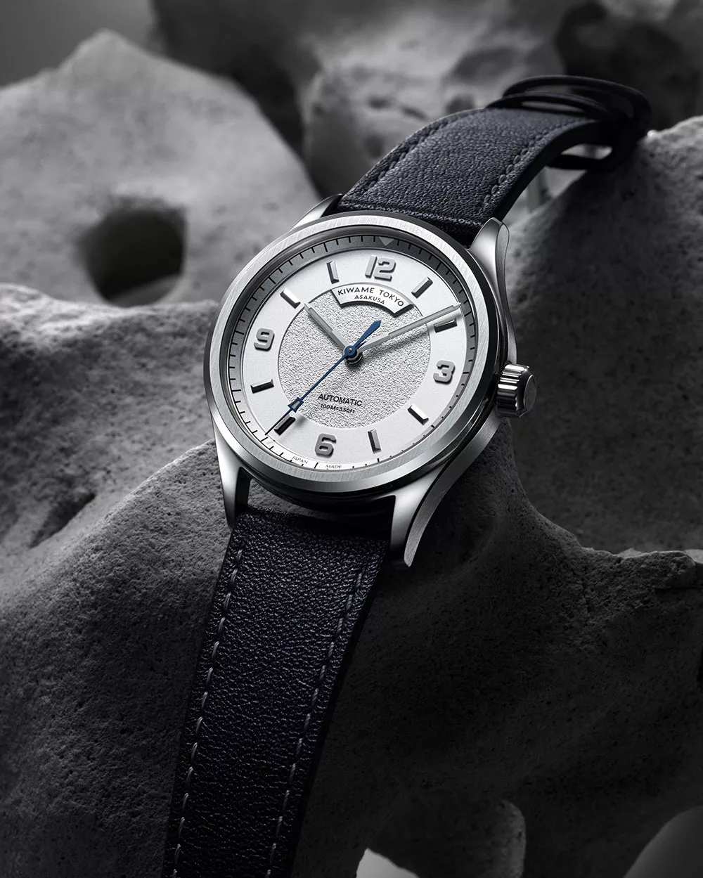 Automatic wristwatch with white textured dial, blue hands, and leather strap displayed on stone background
